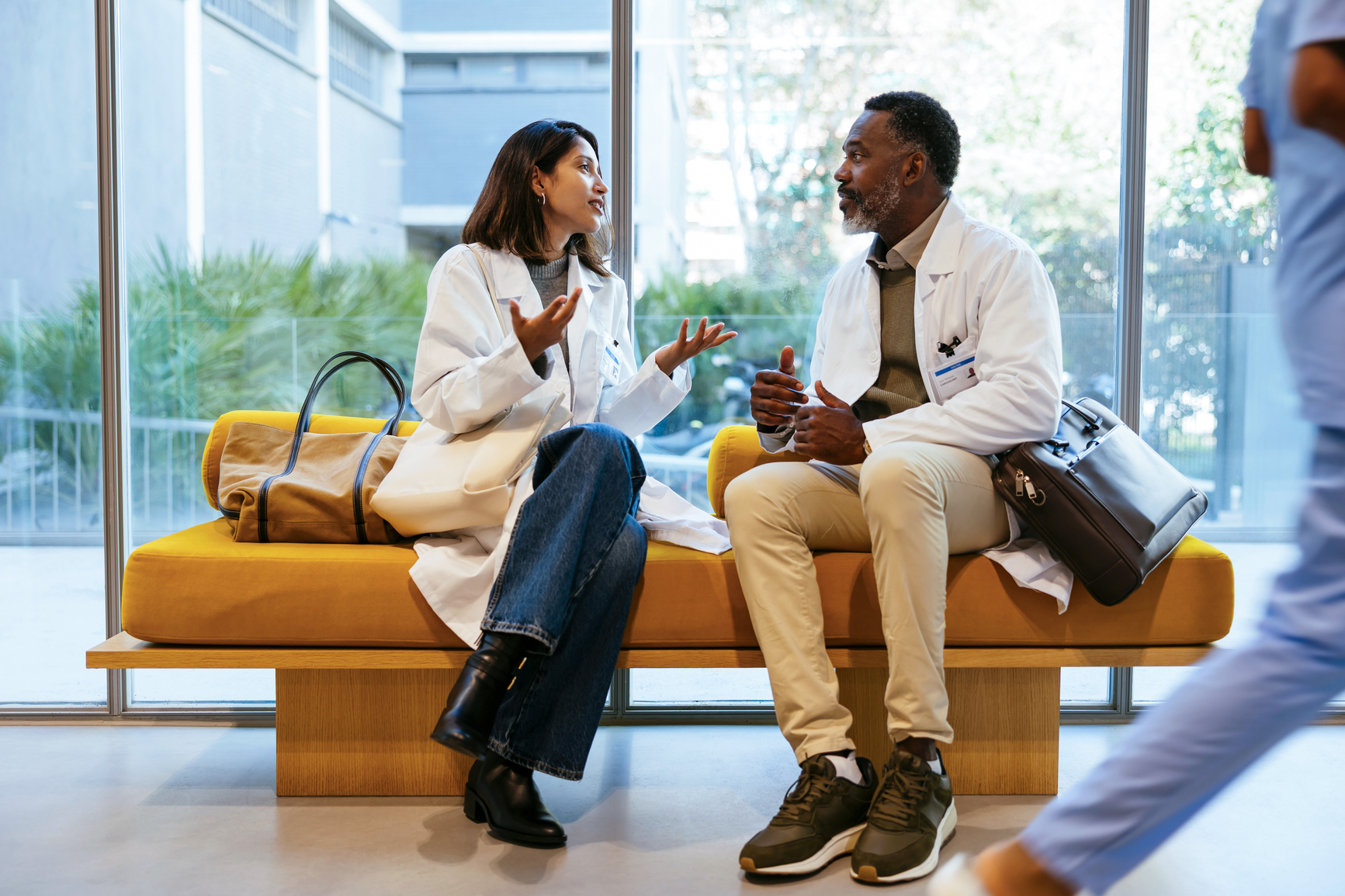 Two medical professionals in conversation, seated on bench in front of windows