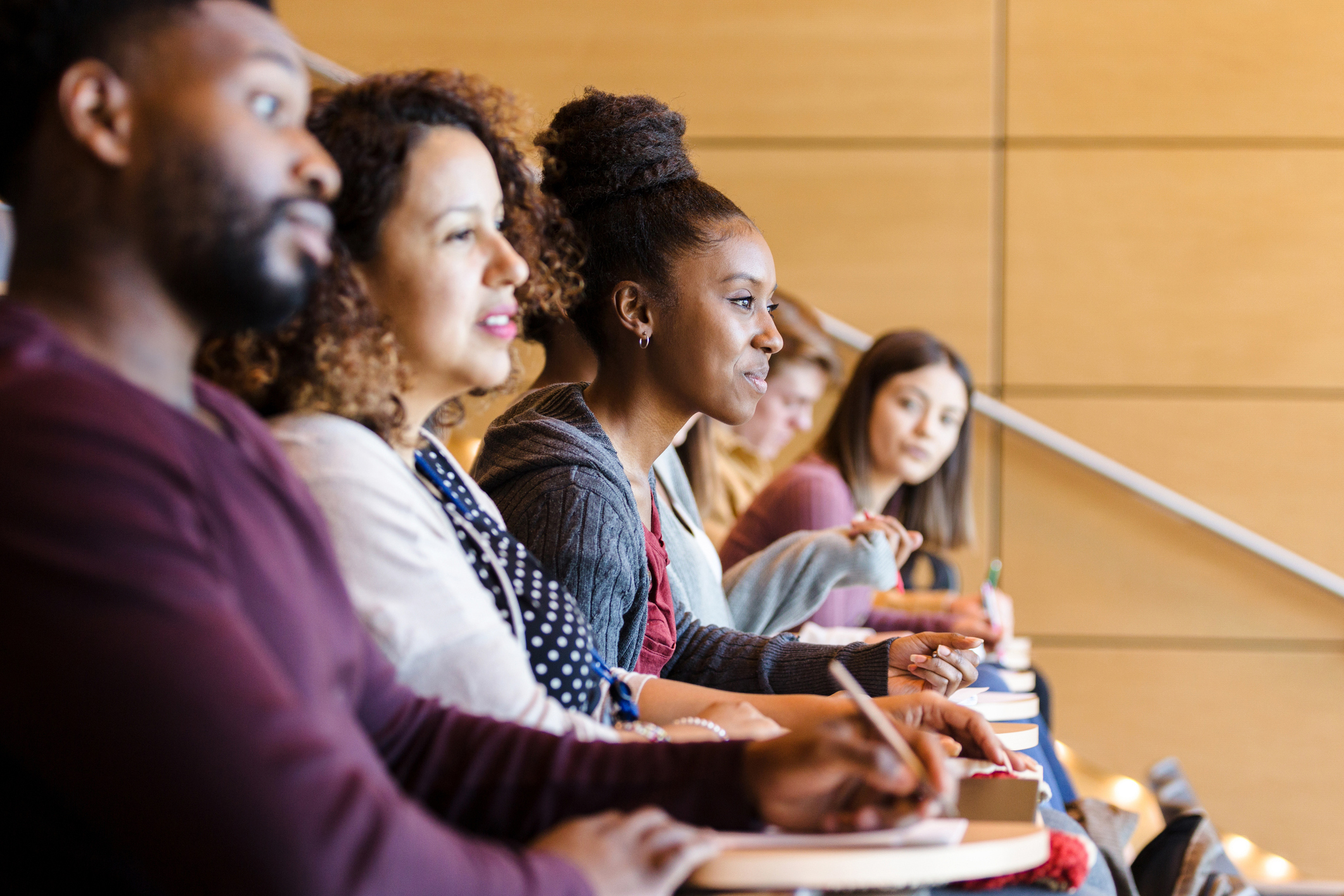 A diverse group of college students are seated in a row in the lecture hall.  Most are engaged in listening to the professor.