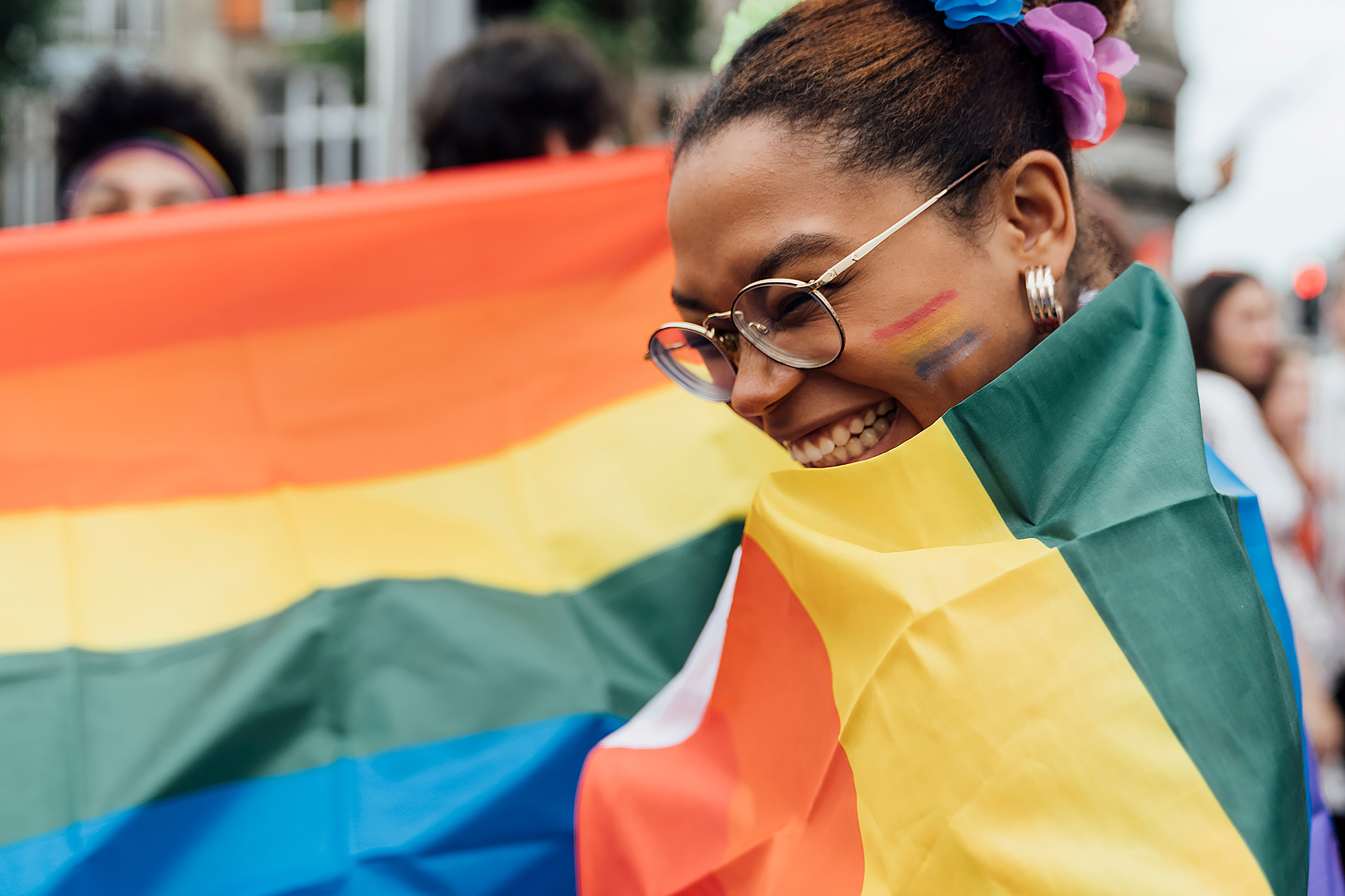 Young Beautiful Black Woman Covered with a Rainbow Pride Flag during the LGBTQ+ Festival in the City. The Street is crowded with Colourful people Enjoying for the event.